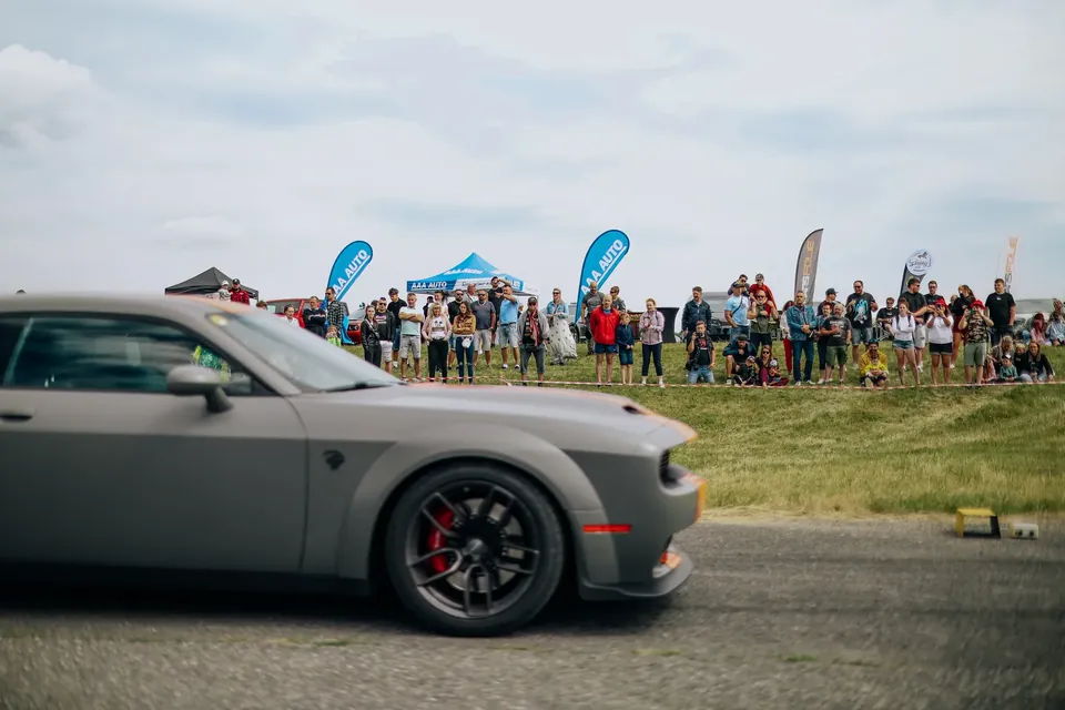 Šedý matný Dodge Challenger projíždí kolem diváků a stanů