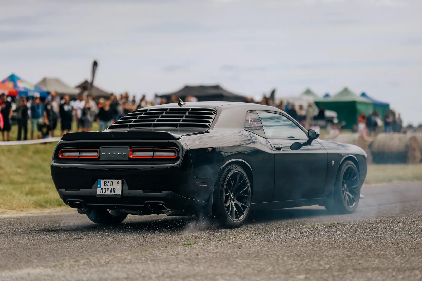 Černý Dodge Challenger dělá burnout na dráze, v pozadí diváci a stany